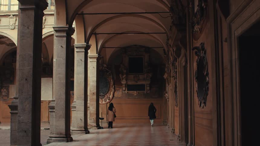 Two people walk through the corridors of a sunlit colonnade with tall columns and ornate walls