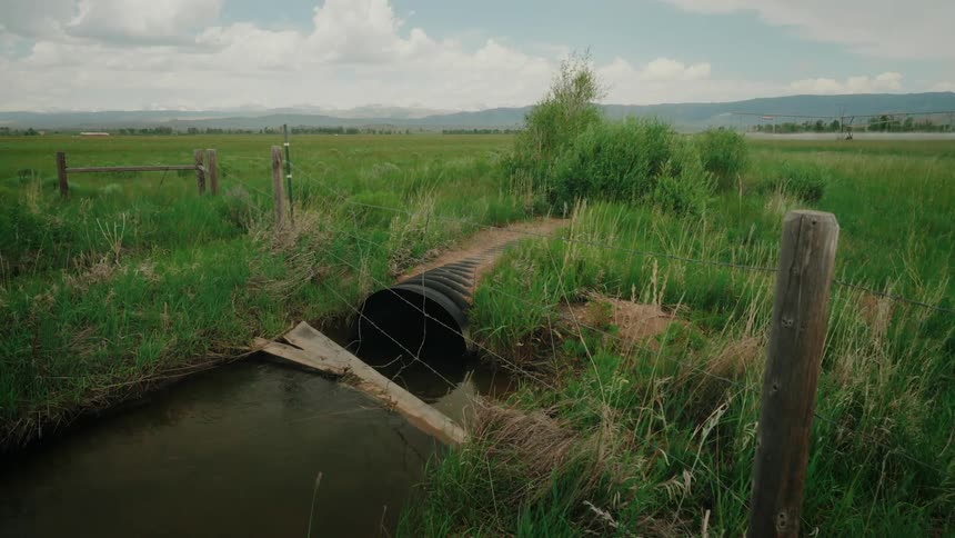 A large culvert pipe carrying water through a grassy field with a wooden fence and a small stream nearby