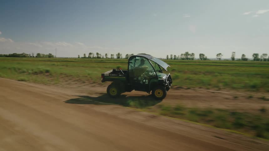 A ranch vehicle with a person and a dog inside driving on a dirt road next to a large grass field