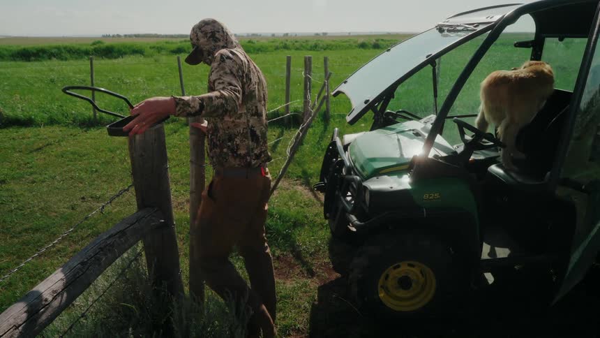 A rancher opens a fence while a dog sits in a green utility vehicle in a grassy field