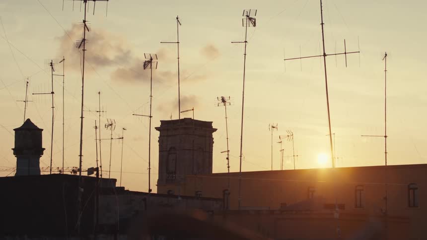 A row of rooftops with numerous antennas silhouetted against a vibrant orange sunset