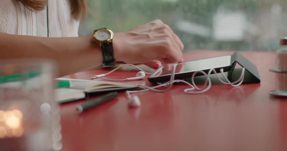 A womans hands using a tablet device while sitting at a table with a cup of coffee and personal belongings