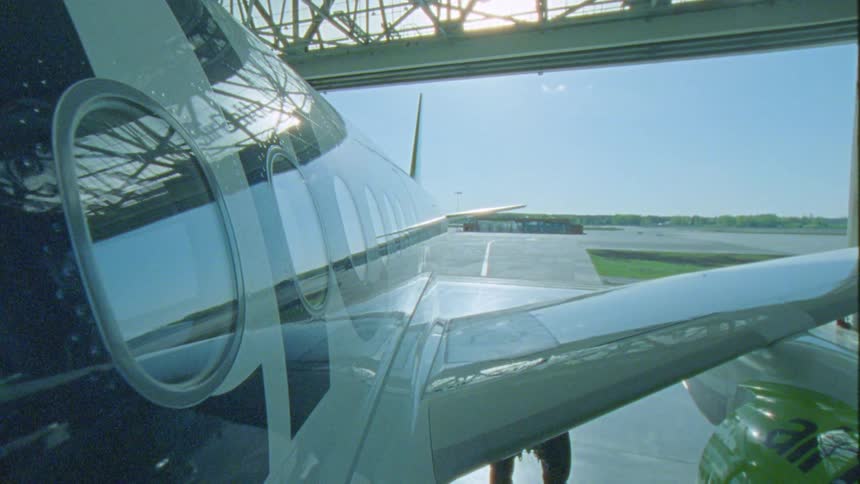 The side of a parked airplane with a glossy reflective surface a row of windows and a wing with a view of the runway visible beyond