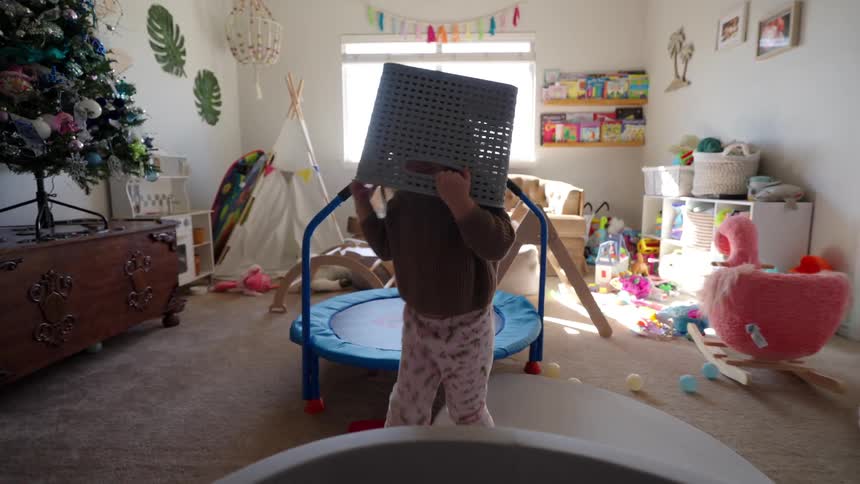 A child is having fun in a room full of toys with a basket on his head next to a blue trampoline and a decorated christmas tree