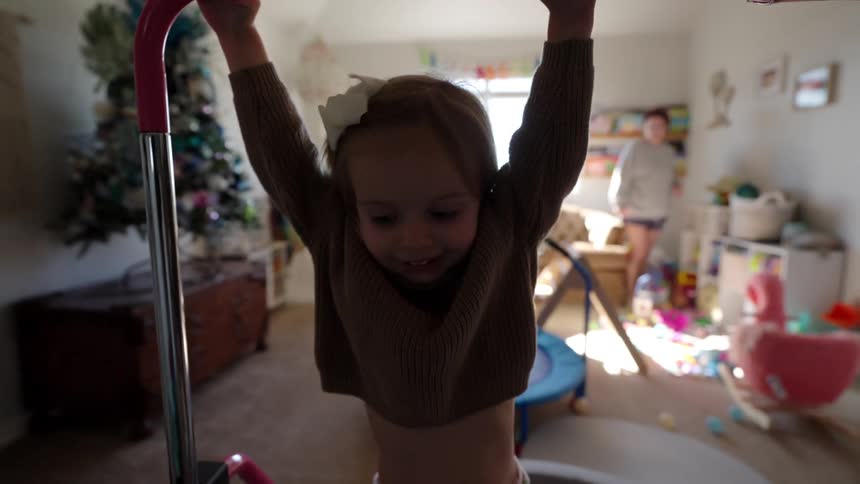 Little girl hangs from a bar while playing while her mother walks across the room in the background
