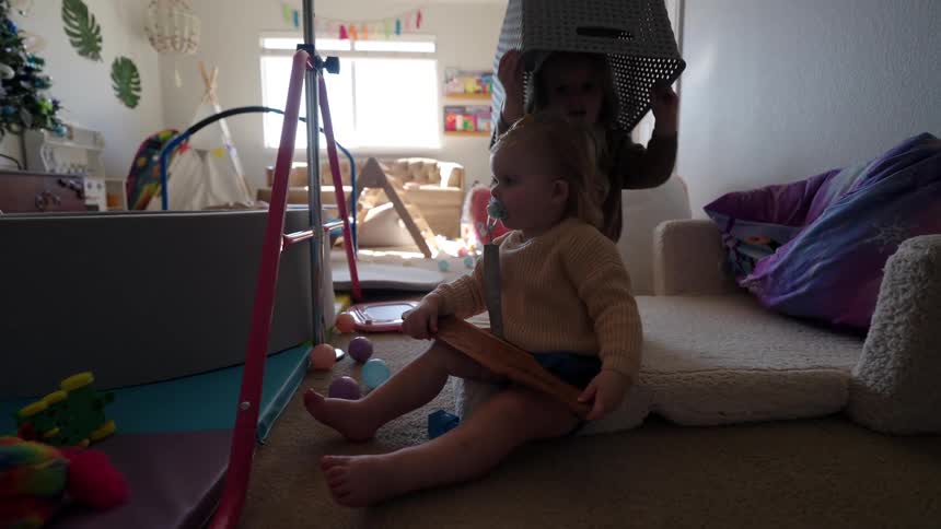 A girl playfully places a basket over the head of her little sister while playing in their bedroom