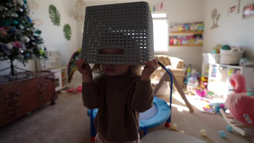 Child with a laundry basket on his head while standing in a room full of toys