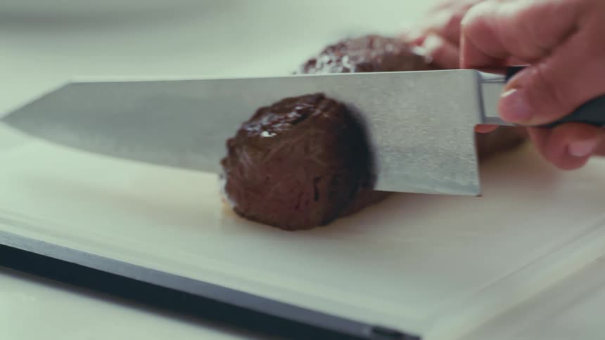 Close up of a hand using a sharp knife to cut a piece of grilled meat on a white cutting board