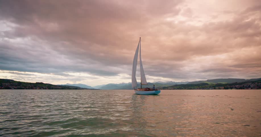 A sailboat glides across a calm lake at sunset with a coastal town nestled among the distant mountains