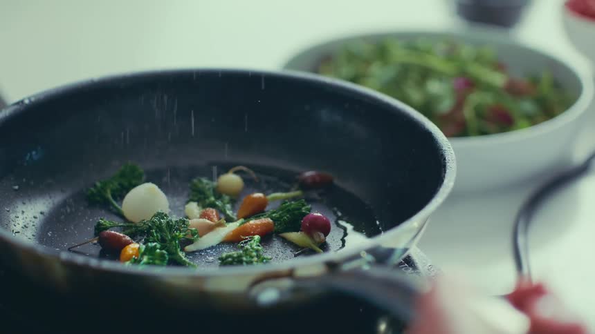 A person sautes tiny carrots and purple onions in a pan with sprinkled salt