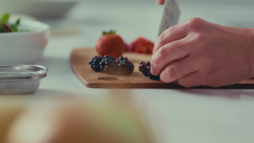 A person carefully slicing a strawberry on a wooden cutting board with other fruits and ingredients visible in the background