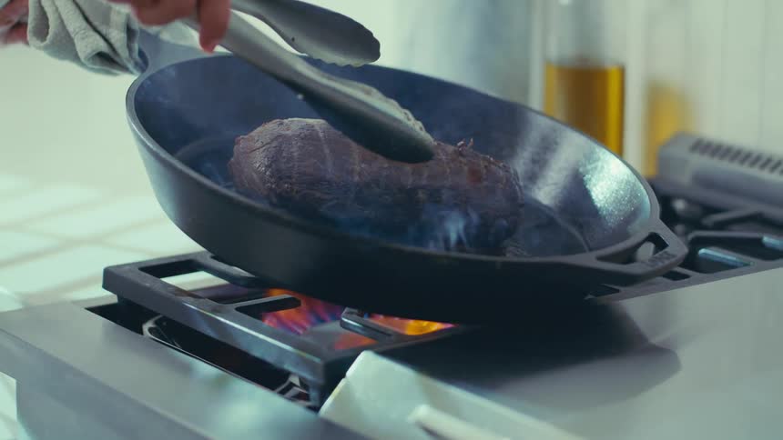 A chef uses tongs to flip a piece of meat in a cast iron skillet