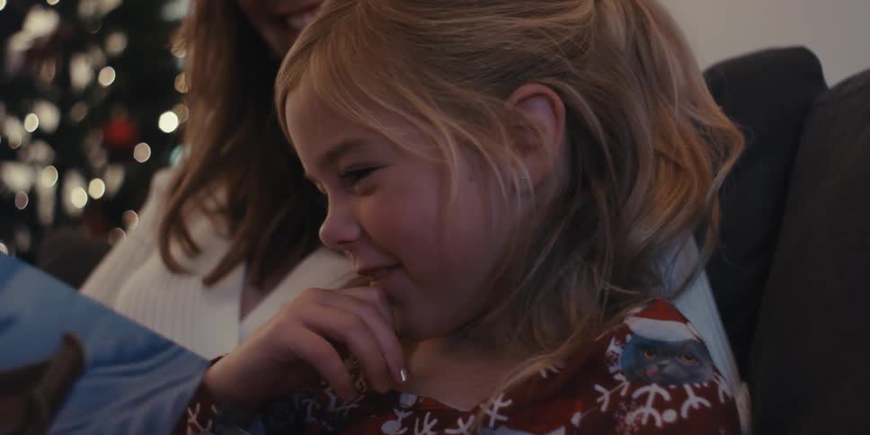 A little girl sits on her mothers lap while they read with a christmas tree in the background