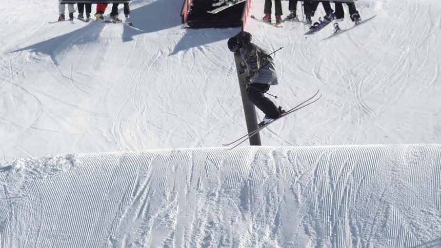 People riding a ski lift while two others perform a trick by siding up a pole and then jumping while on skis