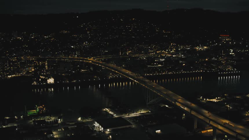 An aerial view of a city illuminated at night showcasing a complex network of highways and overpasses while a bridge adorned with a string of lights gracefully crosses a river winding through the urban landscape