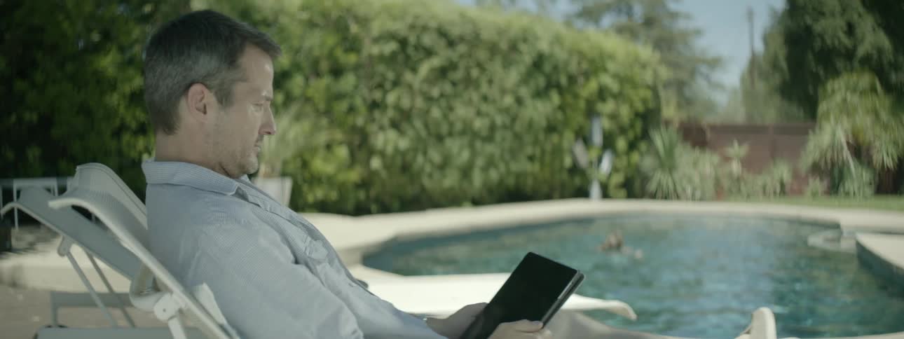A man is reading an ebook while a woman swims in the pool