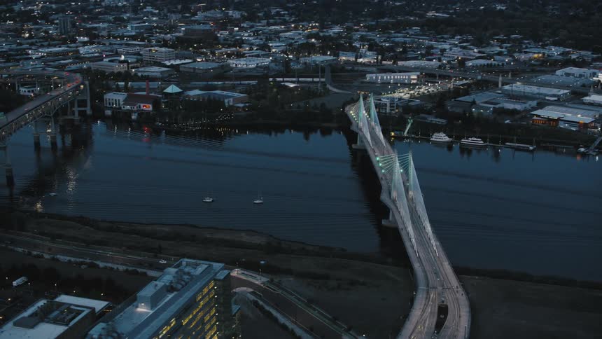An aerial view of portland at dusk shows two bridges spanning the willamette river with traffic moving along them and city lights reflecting on the water