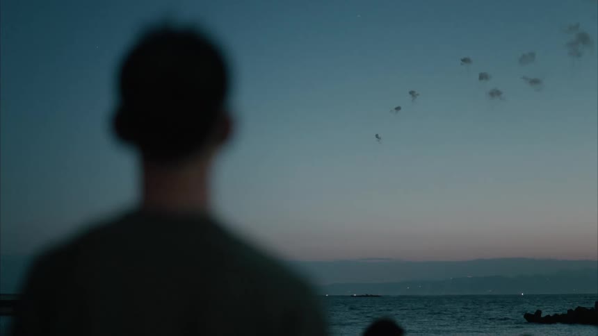 Young man standing along the coast watching fireworks explode over the water at dusk
