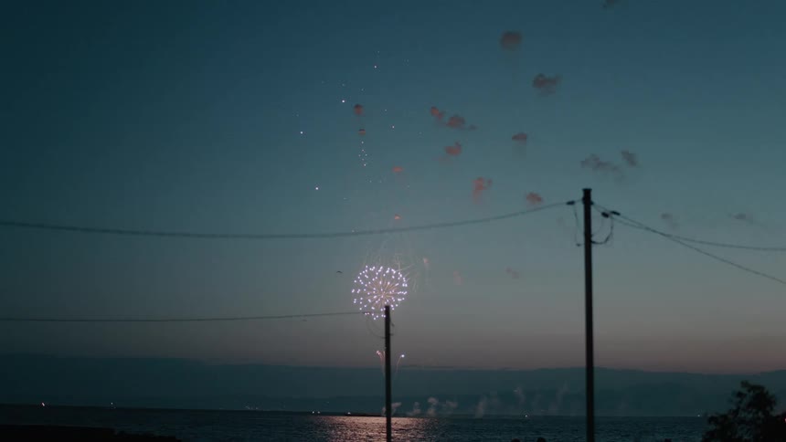 Fireworks glow as they explode over water along the coast at dusk