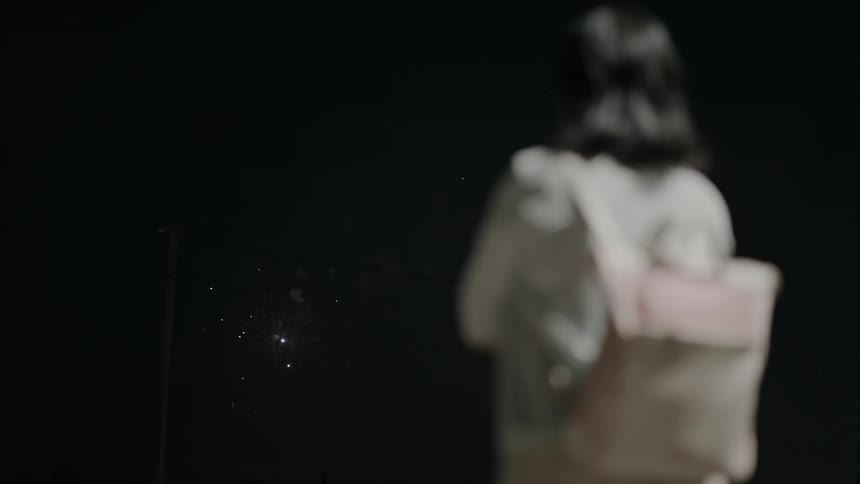 A person with shoulderlength hair and a lightcolored backpack stands outdoors at night watching a display of red fireworks in the sky