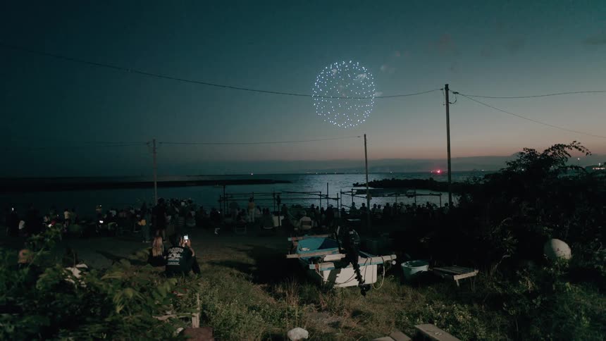 A crowd gathers on a beach at night to watch a fireworks display over the water