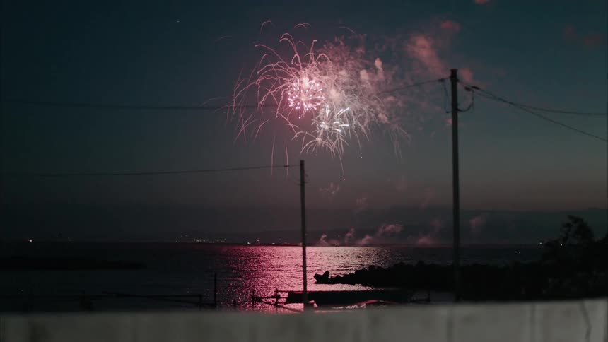 Fireworks fly into the night sky along the coast and glow as they explode over the water