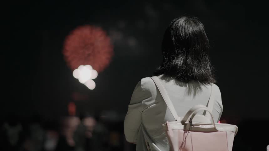 A woman with shoulderlength hair and a lightcolored backpack stands outdoors at night watching a display of red fireworks in the sky