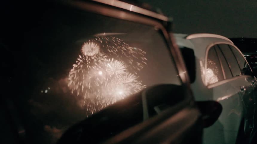 Parked vehicles reflect the fireworks in the sky during an evening fireworks display