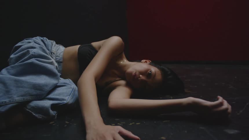 A young woman is lying on a dark floor and staring straight ahead without blinking