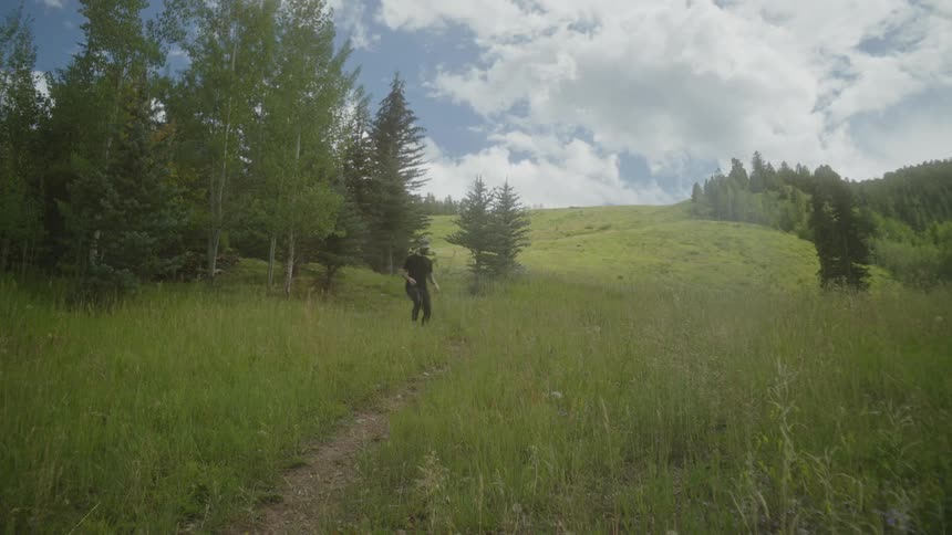 A man in a dark tshirt and cap runs down a dirt path in a grassy field emerging from behind a cluster of trees