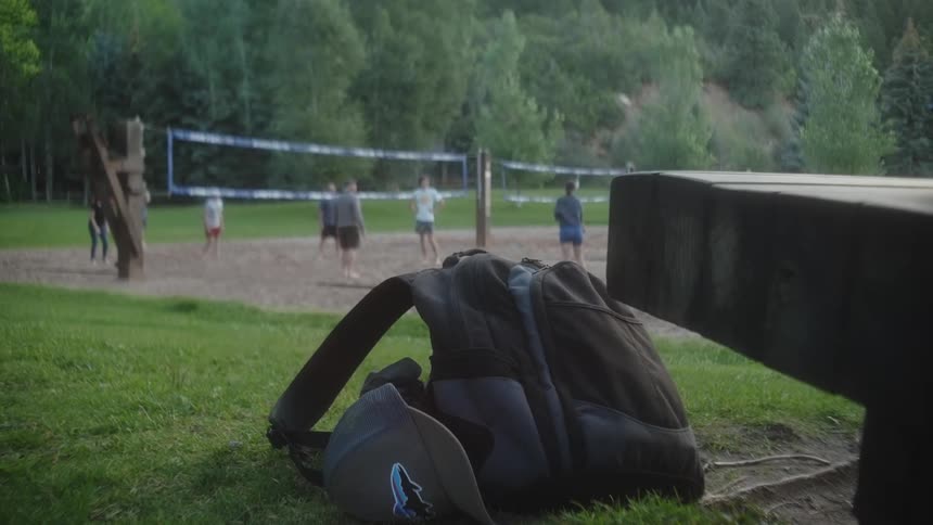 Backpack and cap lie on the grass near a bench while friends play volleyball on a court near a grove of trees