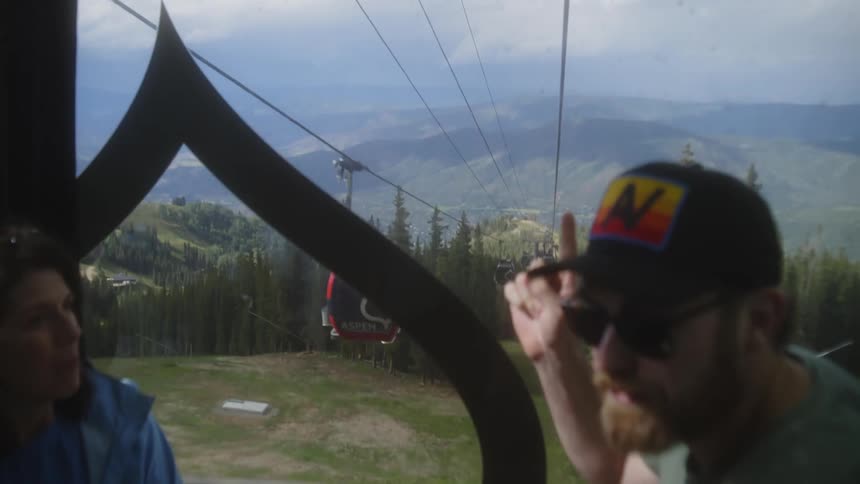 A gondola lift ascends through a forested mountain landscape with distant hills in view while two individuals one wearing a colorful cap are partially visible inside the gondola