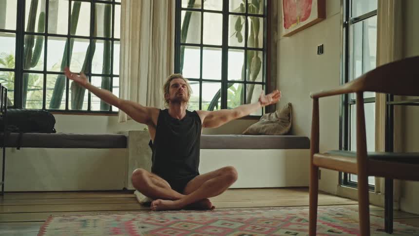 A person is sitting crosslegged on a patterned rug in a welllit room with large windows performing a yoga pose