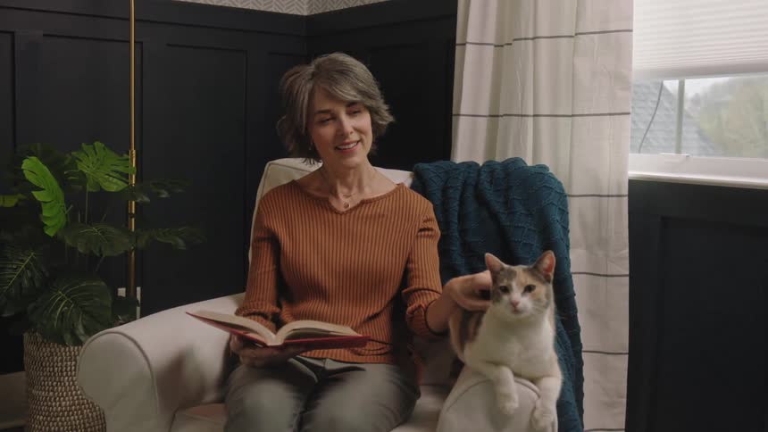 Woman sitting with a book at home smiles while petting her cat