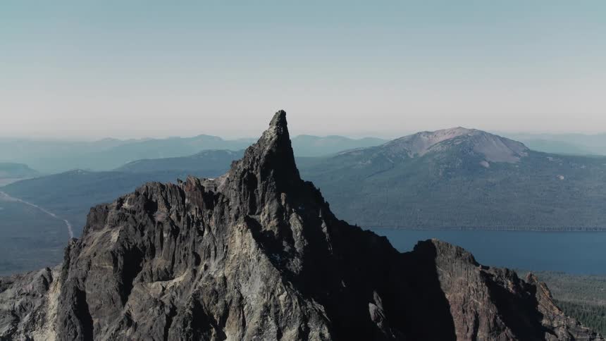 Tall and rough mountain peak overlooking a lake and mountains in the distance under a hazy sky