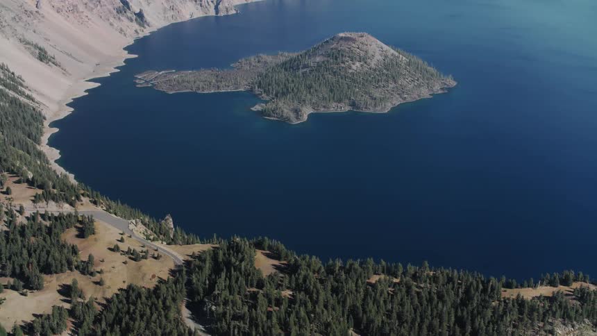 Aerial view of a deep blue lake with an island covered in trees at the center surrounded by a mountainous and forested landscape