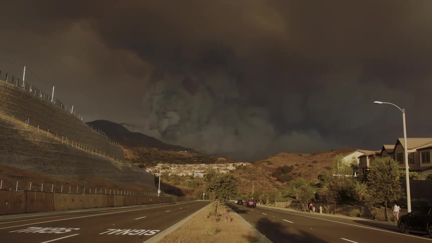 A large plume of smoke rises from a hillside community casting long shadows on the houses and the surrounding landscape as a dark storm cloud gathers overhead