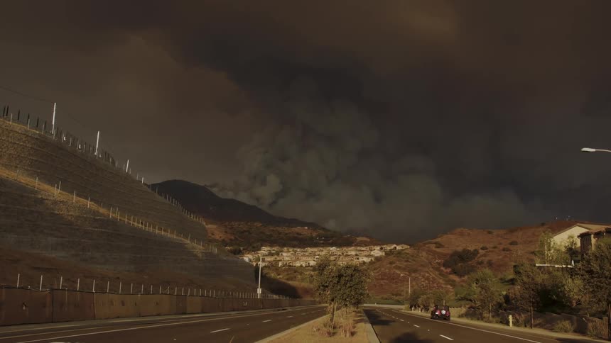 Dark smoke fills the air over a mountainside community as traffic moves on the street