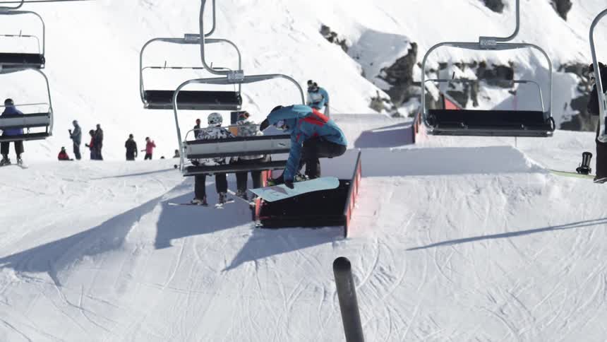 A snowboarder rides a rail on a ski slope with a ski-lift moving overhead