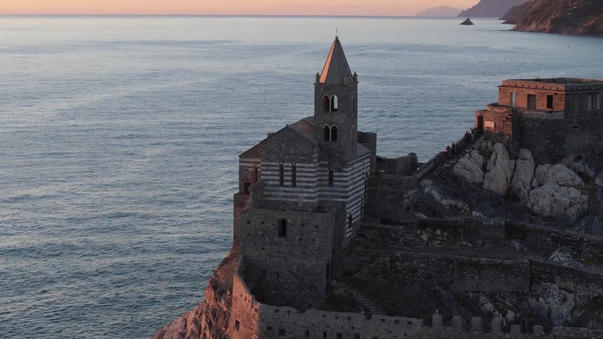 A historic church stands perched on a rocky cliff overlooking the calm blue waters of the mediterranean sea at sunset