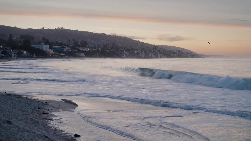 Waves crash onto a sandy beach at dawn with the silhouette of a coastal town visible in the distance