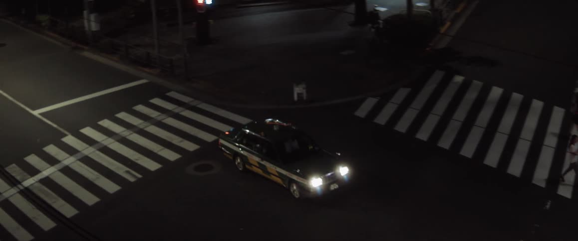 A black and yellow taxi cab drives across a crosswalk at night with a green light its headlights illuminating the street