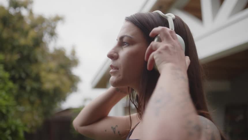 A woman with brown hair tattoos and a black top wears headphones in the backyard