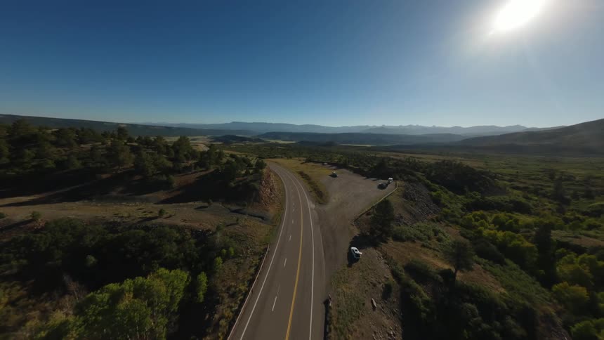 A straight twolane road stretches into the distance through a rugged mountainous landscape with dense green vegetation on either side under a clear blue sky with the sun shining brightly
