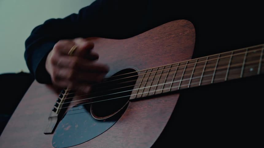 A man with mustache and a black sweater strums the strings of an acoustic guitar