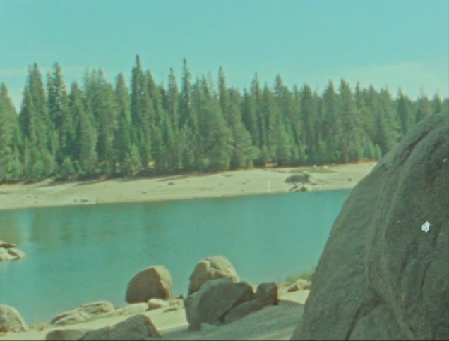 A serene lakeside scene with large boulders in the foreground a calm blue lake in the middle and a dense forest of tall evergreen trees in the background