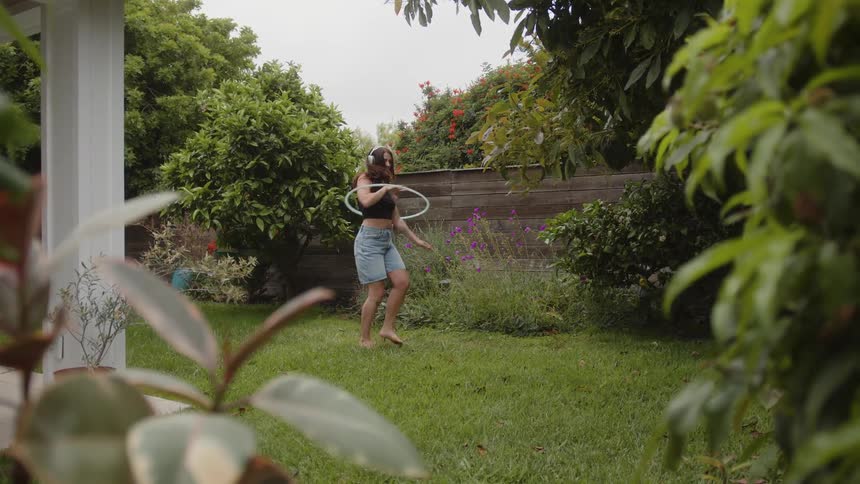 A person wearing a black top denim shorts and headphones is standing on a garden playing with a hula hoop