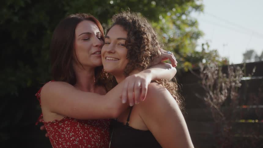 Two smiling women hug and kiss in a yard with a wooden fence on a sunny day
