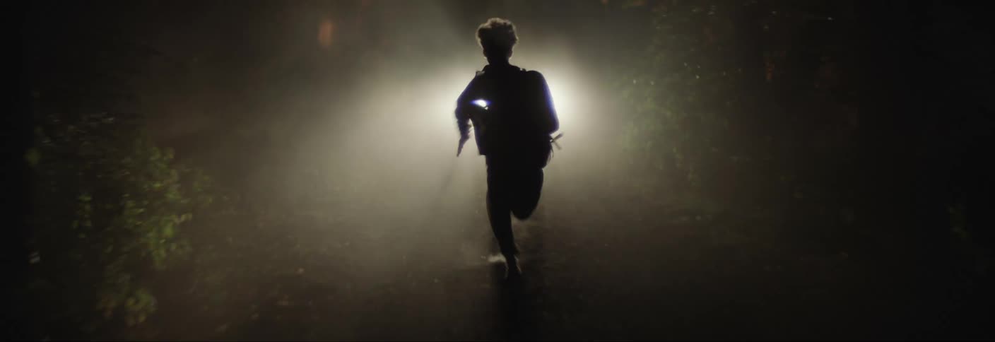 Boy running down a trail at night in a vehicle with headlights on heading towards him