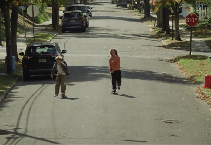 Two boys skateboard down a residential street lined with autumnal trees one performing tricks while the other rides casually